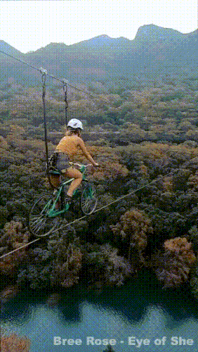woman rides a bike on a high wire over a forest